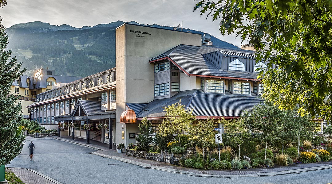 The exterior building and entrance to The Listel Hotel Whistler with a tall mountain backdrop The exterior building and entrance to The Listel Hotel Whistler with a tall mountain backdrop