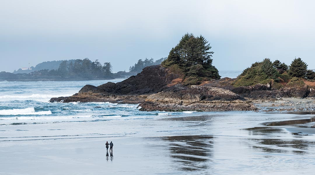 View from Pacific Sands Beach Resort of two people walking along Cox Bay Beach by some rocky terrain in Tofino